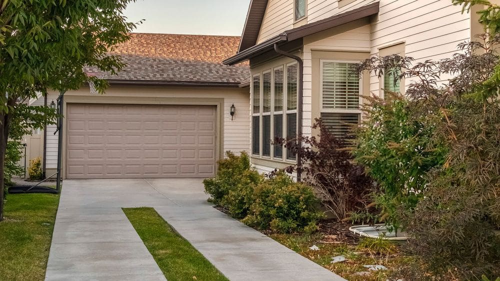 A suburban house in Glendale AZ with beige siding and a brown roof. It features a two-car garage with a closed door. The driveway, showcasing our quality concrete driveway services, has grass growing between the slabs and well-maintained shrubs and small trees on either side. Free quotes available!