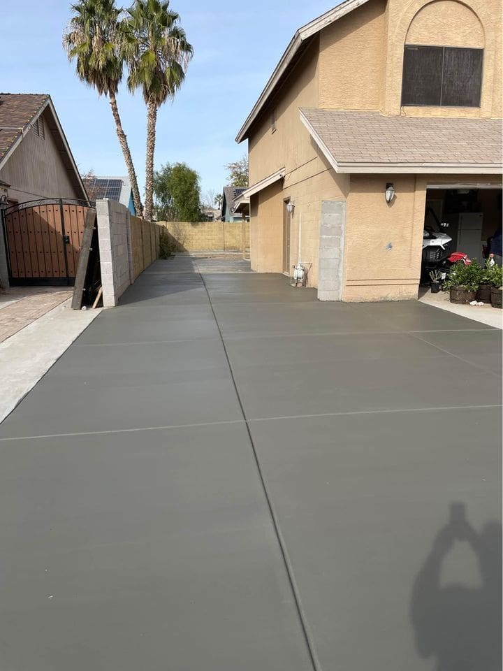 Newly poured by West Valley Concrete, this pristine concrete driveway leads to a two-story beige house with a garage in Glendale, AZ. On the left, a wooden fence and palm trees are visible. A shadow of a person holding a camera is cast on the driveway.