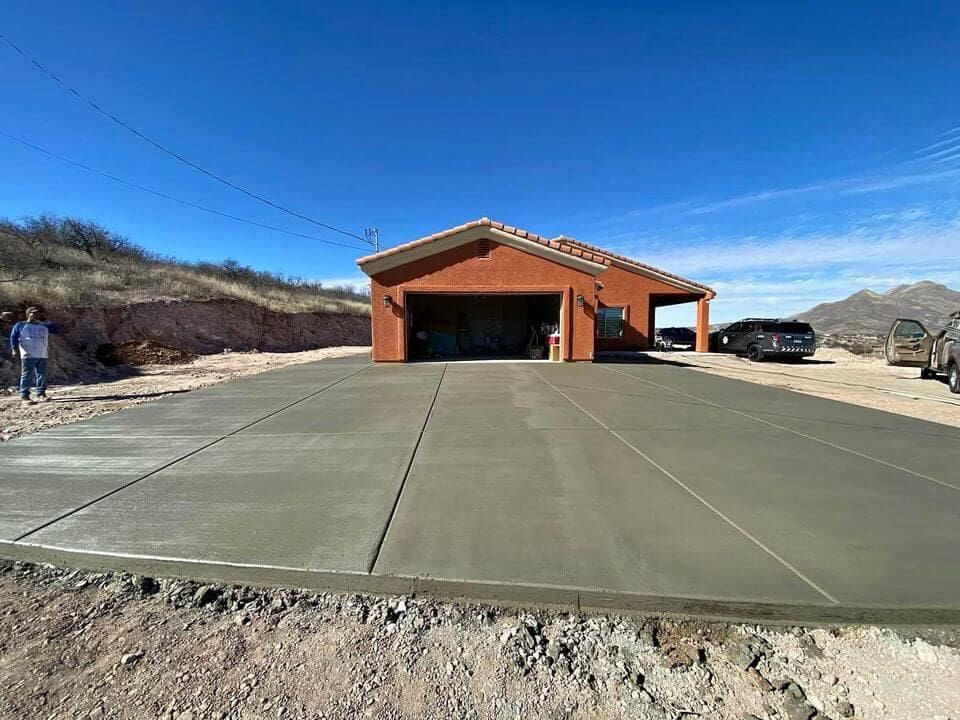 A new concrete driveway crafted by a skilled concrete contractor leads to a brown house with a garage in Goodyear, AZ. Under the clear blue sky and amidst dry terrain, a person stands to the left as a vehicle rests on the right.