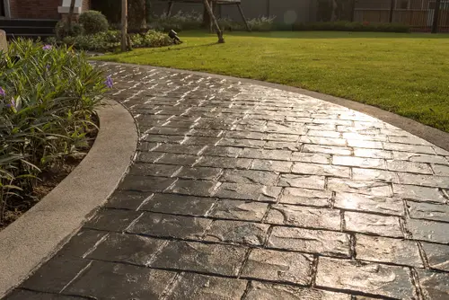 A sunlit path made of textured, rectangular stones curves gently through a garden. Purple flowers bloom along the left side, while a well-manicured lawn stretches beyond the path, reminiscent of the elegance found in concrete patios crafted by skilled contractors in Goodyear, AZ.