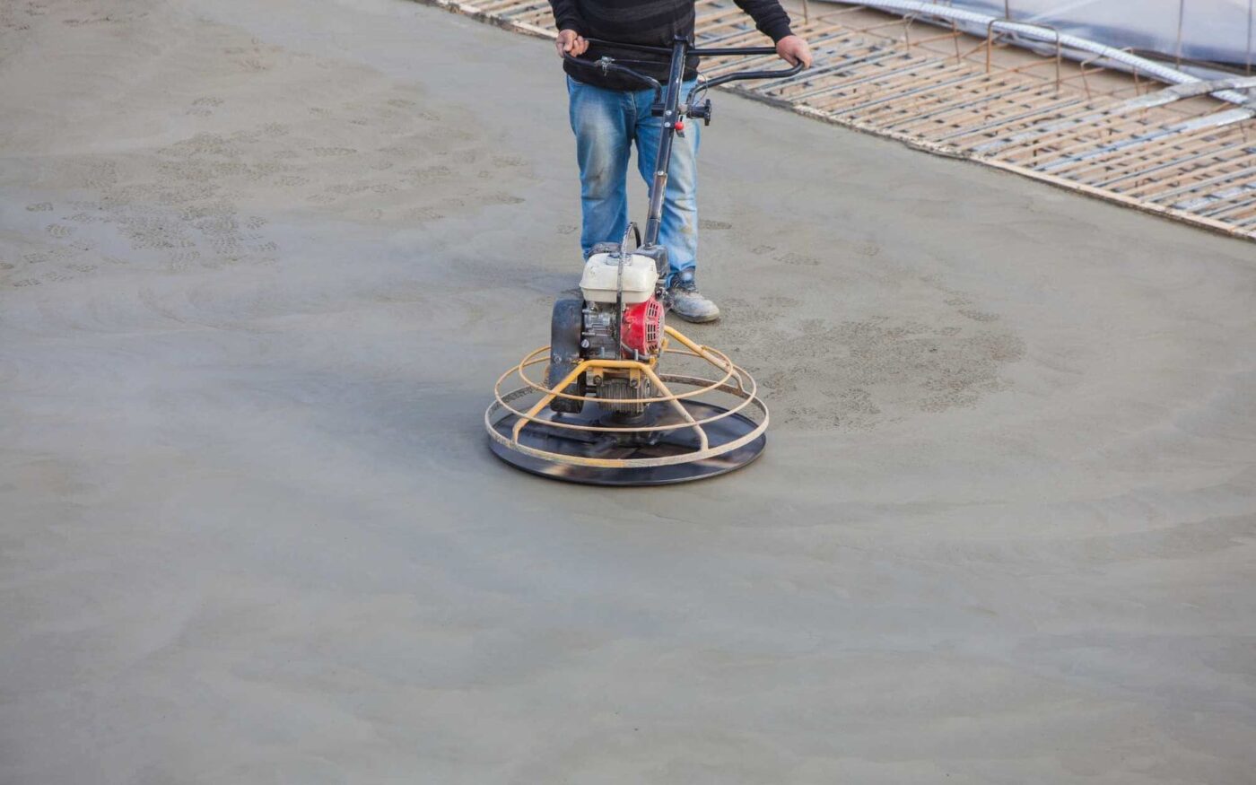 A concrete contractor from Goodyear, AZ, operates a power trowel on a smooth surface, clad in jeans and a dark jacket. The motorized trowel finishes the concrete to perfection, while nearby wooden forms hint at future concrete patios taking shape.