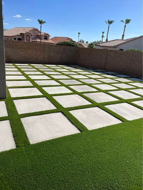 A backyard in Goodyear, AZ boasts a checkerboard pattern of square concrete slabs and lush green grass. Under the clear blue sky, tall palm trees rise while houses nestle in the background, showcasing the artistry of expert concrete patios.