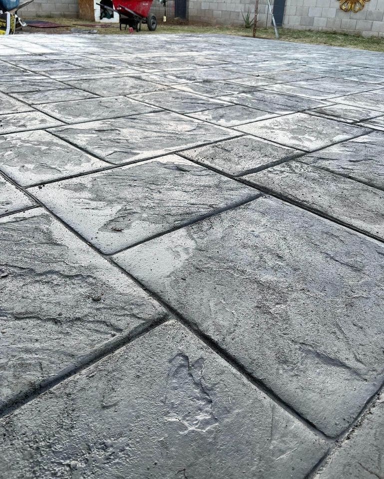 A close-up view of a patterned concrete patio showcases its gray, textured stone-like surface. A red wheelbarrow rests in the background against a garden wall, with greenery at the edges—an ideal project for any skilled concrete contractor.