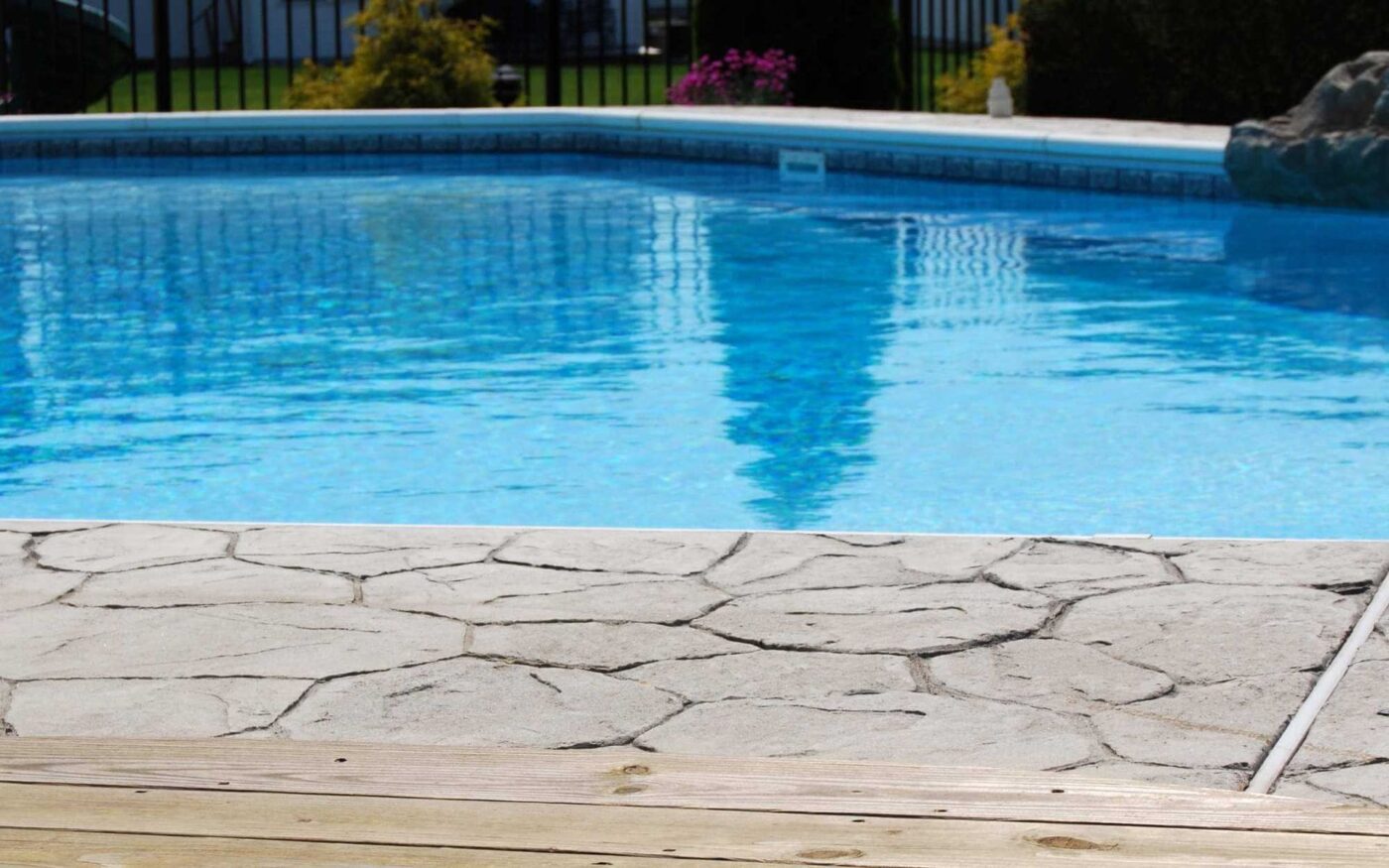 Close-up of a backyard swimming pool with clear blue water in Goodyear, AZ. The foreground showcases a textured stone and wooden deck, expertly crafted by a local concrete contractor. In the background, greenery and fence are partially visible.