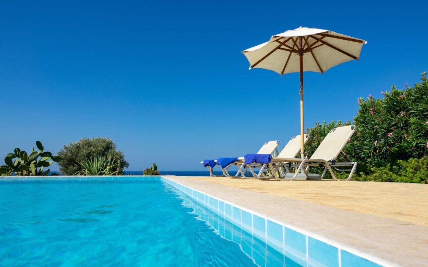 On a sunny day, the swimming pool extends an inviting aura, surrounded by a concrete patio where three lounge chairs and a large umbrella stand. The clear water mirrors the blue sky as lush greenery borders the scene. Towels rest neatly on two chairs, enhancing this serene oasis.